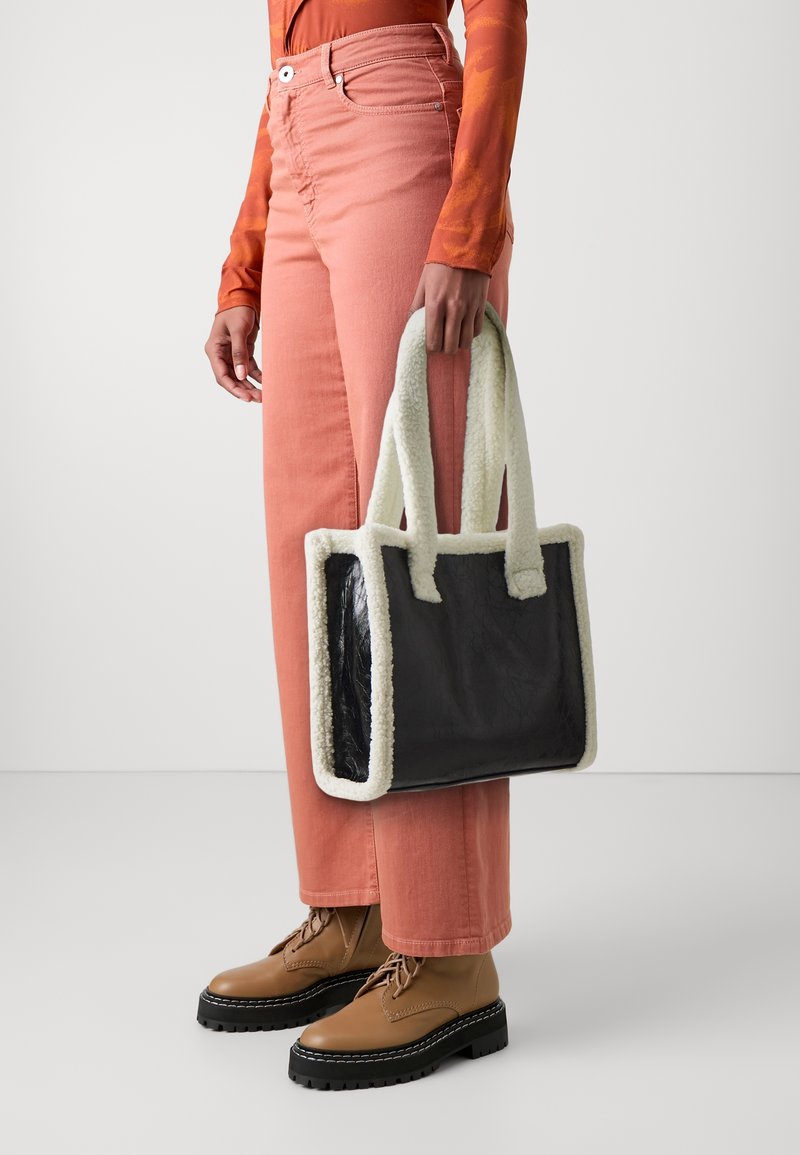 Personne portant un haut orange et un pantalon rose tenant un sac à main rectangulaire noir avec une bordure en polaire blanche et des bottes marron à lacets avec semelles épaisses.