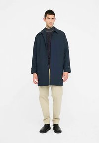 Tamsiai mėlynas tranšėjos paltas, pagamintas iš lengvos medžiagos, su apykakle, sagomis ir šoninėmis kišenėmis, dėvimas ant riboto megztinio ir smėlio spalvos kelnių.