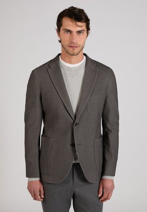 Man draagt een grijze textuurblazer over een lichtgrijze trui en een wit onderhemd, staand tegen een effen lichte achtergrond.