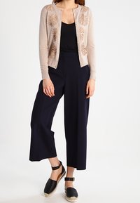 Cardigan beige con ricami floreali, canotta nera, pantaloni a gamba larga blu navy e scarpe espadrilles nere con cinturino alla caviglia.
