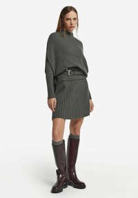 Maglione grigio a coste con collo alto e orlo asimmetrico abbinato a una gonna a coste. Stivali bordeaux al ginocchio con accenti grigi.