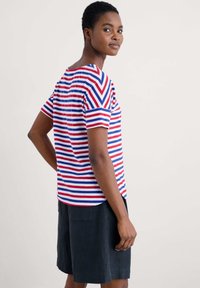 Striped short-sleeve shirt in white, red, and blue. Features a relaxed fit with a rounded neckline and side slits. Paired with black shorts.