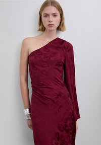 Robe bordeaux à une épaule en tissu texturé avec un motif subtil, présentant un design drapé et des manches longues. Accent avec un bracelet argenté.