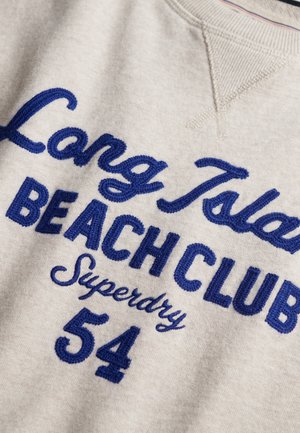Pull beige avec texte brodé en bleu disant "Long Island Beach Club Superdry 54" sur le devant.