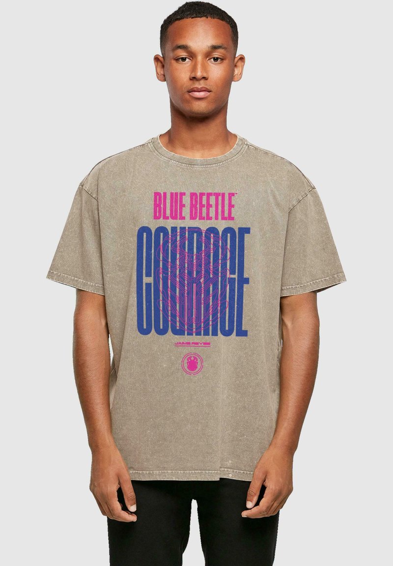 Camiseta holgada de color marrón claro con un estampado gráfico que presenta "BLUE BEETLE" en rosa y "COURAGE" en azul. Cuello redondo y mangas cortas.