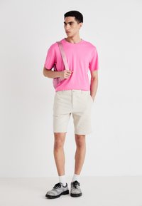 Šviesiai rožinė marškinėliai, kreminės šortai, juodi ir pilki batai, balti kojinės, laiko šviesiai rožinę rankinę, stovi ant balto fono.