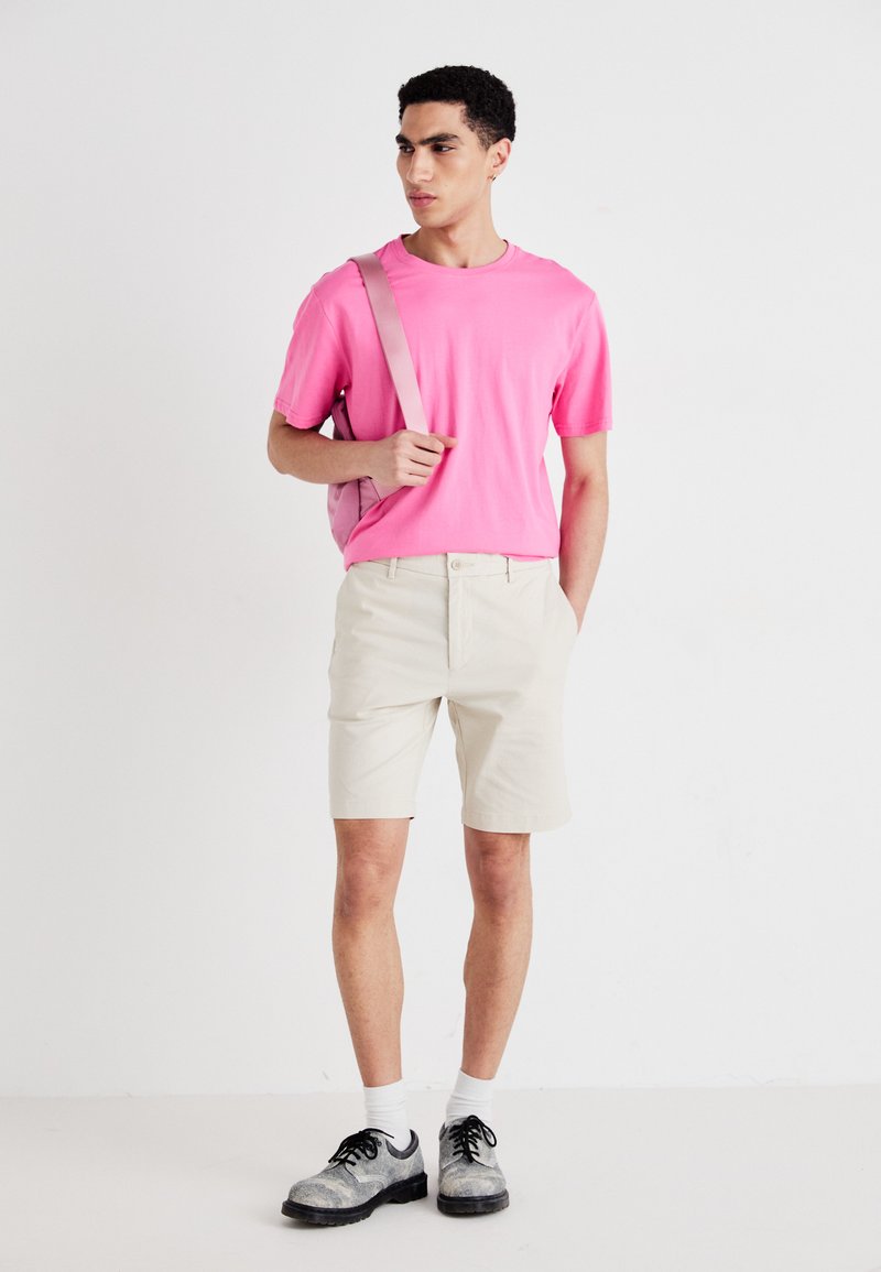 Šviesiai rožinė marškinėliai, kreminės šortai, juodi ir pilki batai, balti kojinės, laiko šviesiai rožinę rankinę, stovi ant balto fono.