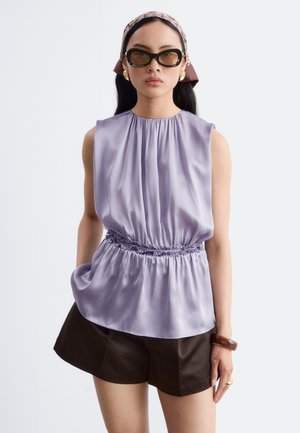 Femme portant un chemisier en satin lavande sans manches et un short marron foncé, avec un foulard à motifs, des lunettes de soleil foncées, des boucles d'oreilles en or et un bracelet.