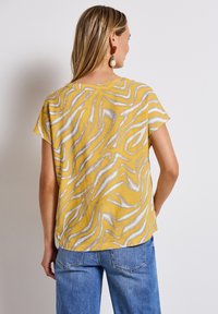 Top giallo a maniche corte con un motivo ondulato bianco e grigio, realizzato in tessuto leggero. Taglio con vestibilità rilassata e scollo rotondo.