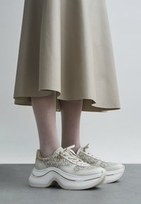 Bele superge z vzorcem teksture in zlatimi poudarki, z debelim podplatom in vezalkami, nošene z bež krilom.