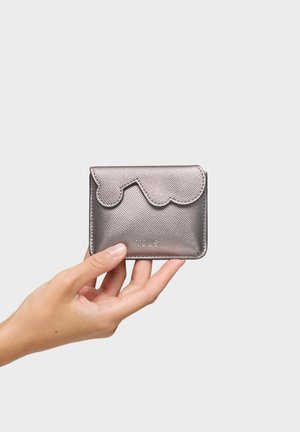 Mano sosteniendo una pequeña cartera de cuero plateado metálico con solapa con borde ondulado y nombre de la marca en relieve en el frente sobre un fondo liso.