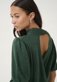 Zielony, błyszczący sweter z bufiastymi rękawami i wycięciem z tyłu. Materiał ma szeleszczącą teksturę, podkreślającą dopasowany krój.