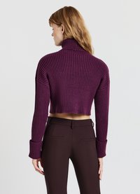 Kopfnuss, gerippter Rollkragenpullover in tiefem Lila. Lange Ärmel mit breiten Bündchen. Kombiniert mit dunkelbraunen, maßgeschneiderten Hosen.
