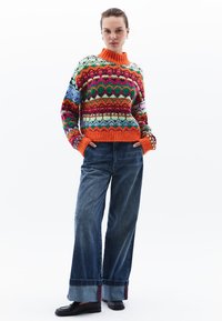 Kolorowy sweter dzianinowy z wielokolorowymi wzorami i pomarańczowym wysokim kołnierzem, w połączeniu z jeansami o szerokich nogawkach z podwiniętymi brzegami.