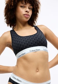 Sort sport bh med et geometrisk mønster, bred hvid elastik med "Calvin Klein" logo. Blødt stof, scoop neck design, og uden polstring.