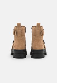 Botas de tobillo de ante en tono beige con suela de goma negra, con cremalleras laterales y lazos de tela para poner y quitar fácilmente. Diseño de punta cuadrada.