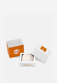 Boîte blanche avec un couvercle orange, portant le logo Timberland. À l'intérieur, un article beige partiellement visible, avec une surface en tissu texturé.