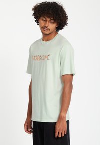 Ljusgrön bomullströja med korta ärmar, med en texturerad orange "VOLCOM" logotyp på framsidan. Enkel, avslappnad passform.