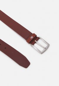 Ceinture en cuir marron avec une boucle en métal argenté, au design plat, présentant un patch logo rouge et bleu près de la boucle et une couture soignée.
