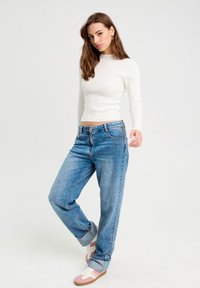 Haut à manches longues blanc à côtes associé à un jean bleu clair à coupe décontractée, comportant une coupe droite et des poignets retroussés. Les chaussures ont des accents roses.