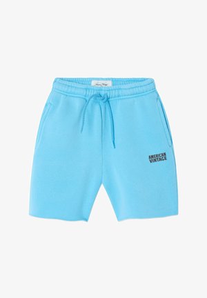 Osoba nosi svijetloplave kratke hlače od flisa s natpisom "American Vintage", smeđu majicu dugih rukava i bež klompe s ukrasnim privjescima.