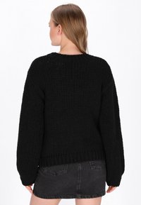 Schwarzer Strickpullover mit lockerer Passform, weiten Ärmeln, gerippter Textur und rundem Halsausschnitt. getragen über einem schwarzen Jeansrock.