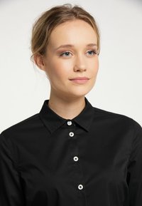 Młoda kobieta o jasnej skórze i blond włosach związanych z tyłu, nosząca czarną koszulę zapinaną na guziki, patrząca lekko w bok z neutralnym wyrazem twarzy.