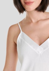 Camiseta blanca sin mangas con un diseño de escote en V, tirantes finos y tejido texturizado. La parte superior muestra un ligero brillo y una estética minimalista.