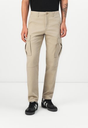 Beige cargo pants with multiple side pockets, straight legs, and a textured fabric, paired with black athletic shoes.