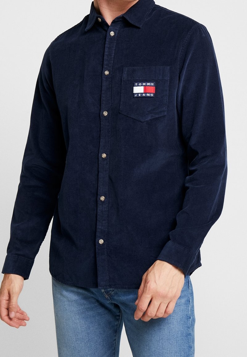 Marineblauwe corduroy overhemd met lange mouwen, knopenlijst, kraag en een borstzak met een logo patch in rood, wit en blauw.