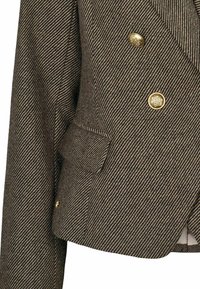 Gros plan d’un blazer marron à motif chevrons avec des boutons décoratifs dorés et une petite poche à rabat au niveau de la poitrine.