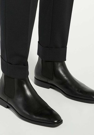 Black leather chelsea boots with elastic side panels, pointed toe design, and a smooth texture, paired with tailored black trousers.