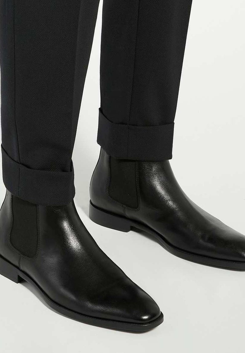 Zwarte leren chelsea-boots met elastische zijkanten, puntige neus en een gladde textuur, gecombineerd met op maat gemaakte zwarte broeken.