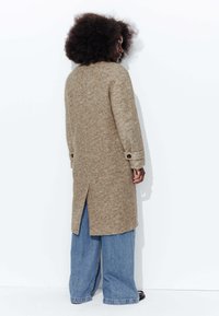Manteau beige en chevrons avec de longues manches, poignets à un seul bouton et fente au dos, associé à un pantalon en denim bleu clair ample.