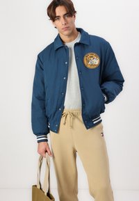 Mørkeblå bomberjakke med logo-patch, stripete mansjetter, brukt over en grå sweatshirt og beige joggebukser, holder en lys brun totebag.