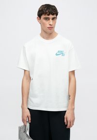 Άσπρο βαμβακερό T-shirt με κοντά μανίκια, που διαθέτει λογότυπο Nike SB σε ανοιχτό μπλε χρώμα στην αριστερή πλευρά του στήθους. Το μοντέλο φοράει μαύρο παντελόνι και κρατά μια τσάντα.