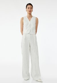 Biały top bez rękawów w pasy z dekoltem w kształcie litery V, pięcioma guzikami i dopasowanymi spodniami w szerokiej nogawce. Materiał jest gładki z granatowymi paskami.