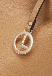 Sac en simili cuir texturé marron, doté d'une étiquette ronde en or avec "Credi Munch" en relief et d'une attache de sangle noire.