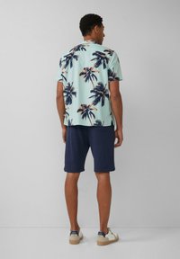 Korte mouwen, lichtblauw shirt met donkere palmbomenprints, gecombineerd met marineblauwe shorts. De outfit heeft een relaxte pasvorm en casual sneakers.
