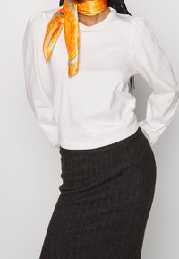 Camicia bianca a maniche lunghe con maniche a sbuffo abbinata a una gonna a matita nera a coste. Sciarpa di seta arancione con dettagli decorati legata attorno al collo.