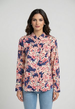 Donna che indossa una camicia a maniche lunghe con bottoni, decorata con un motivo floreale rosa e blu navy, abbinata a jeans azzurri, in piedi davanti a uno sfondo grigio.