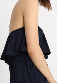 Námořnická modrá off-shoulder blůza s volánkovou vrstvou, elastickým pasem a měkkou texturou, navržená pro pohodlné nošení.