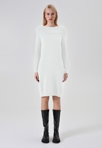 Abito lavorato a maglia bianco con ampio scollo a barca e maniche lunghe, caratterizzato da una vestibilità aderente e che arriva a metà polpaccio, abbinato a stivali neri fino al ginocchio.