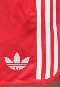 Κόκκινα αθλητικά σορτς με λευκό λογότυπο τριφυλλιού Adidas και τρεις κατακόρυφες λευκές ρίγες κατά μήκος της πλαϊνής ραφής.