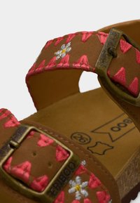 Καφέ σανδάλι με ροζ και λευκή κεντητή floral διακόσμηση, δύο ορειχάλκινα αγκράφες και μαξιλαρωτή σόλα που δείχνει το μέγεθος και τα εικονίδια δέρματος.
