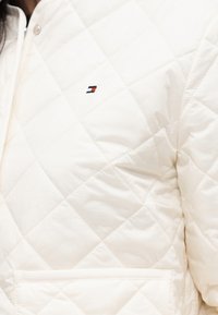 Veste blanche matelassée avec couture en losanges, poche latérale et petit logo rouge, blanc et bleu sur la poitrine.