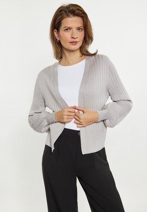 Lichtgrijze geribbelde cardigan met een v-hals, brede mouwen en een zoom die aan de zijkanten licht omhoog loopt. Gepaarde met een witte top en zwarte broek.