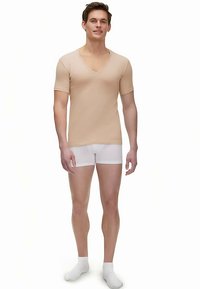 T-shirt beige à manches courtes avec col en V, fabriqué en tissu extensible, associé à un boxer blanc et des chaussettes blanches, porté par un modèle masculin debout.