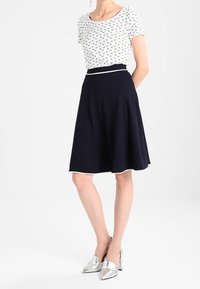 Top bianco con motivi blu navy, abbinato a una gonna blu navy con vita aderente e orlo svasato. Scarpe argentate completano il look.