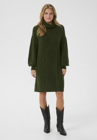 Sieviete, kas ģērbusies tumši zaļā adītiem rakstiem turtleneck kleitā ar pūkveida piedurknēm un melnām ādas kedām līdz pusceļam, stāv uz balta fona.